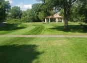 Thumbnail 11 of 54 - Large green pasture with a sidewalk across the middle. Gazebo with tables and chairs can be seen by the trees surrounding the grass. Blue and cloudy sky.