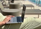 Thumbnail 8 of 21 - A person is using a laptop on a lounge chair by a pool.