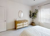 Thumbnail 17 of 21 - A white bedroom with a bed, dresser, mirror, and plant.