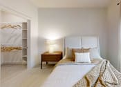Thumbnail 19 of 21 - A bedroom with a bed, nightstand, and window with blinds.