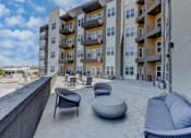 Thumbnail 25 of 51 - A modern apartment complex with a rooftop patio furnished with a couch, chairs, and ottoman.