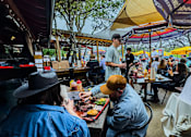 Thumbnail 42 of 46 - A man and a woman are sitting at a food stand with a man standing behind them.