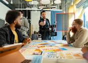 Thumbnail 7 of 15 - A group of people are sitting around a table with papers and pens, one person is standing and gesturing with their hands.