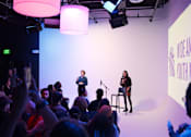 Thumbnail 14 of 15 - Two women are speaking to an audience in a dimly lit room.