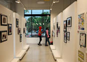 Thumbnail 13 of 15 - Two people are walking through an art gallery with white walls and pictures on the walls.