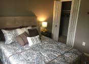 Thumbnail 11 of 14 - Bedroom with tan walls and a tan headboard. The bed is made in a blue patterned bed spread and white and brow accent pillows. Next to the bed is a lamp on a side table. The white closets doors are open to the right of the bed.