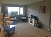 Thumbnail 9 of 14 - Living room with tan carpet and grey walls. There is a large pictured window on the back wall with two chairs under it. There is a couch against the left wall with a lamp and table next to it. Across from the couch on the right wall is a entertainment center. Art is also hanging on the right wall.
