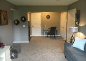 Thumbnail 10 of 14 - Living room with tan carpet and grey walls. A couch is up against the right wall. In the distance is a table and chair.
