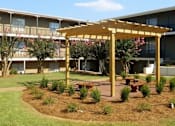 Thumbnail 7 of 47 - A wooden pergola with a bench underneath it is in the foreground of a building.