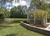 Thumbnail 9 of 16 - White gazebo to the right of photo with trees, greenery and lined with wood surround.  Green grass in the middle leading up to a pond with a fountain.  Sky and green trees in the background.