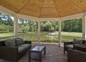 Thumbnail 10 of 16 - Interior of gazebo with with light wood ceiling and white trim. Wood slatted surround with brick flooring.  Green couch with coffee table to the left, and two green/brown chairs to the right.