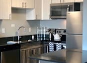 Thumbnail 20 of 47 - Image of a kitchen in the Jefferson. There are dark brown wood-like cabinets below and white cabinets on top. There are black countertops with stainless steel appliances. There is a sink to the left with a dishwasher and a stove and fridge on the right. The walls are a light grey with wood-like flooring.