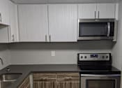 Thumbnail 19 of 47 - Image of kitchen, there are dark wood-like cabinets below with white cabinets on top. There are black countertops and stainless steel appliances. There is a sink to the left and an oven with a microwave above on the right. The walls are a light grey.