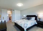 Thumbnail 18 of 47 - Photo of a bedroom at The Jefferson. The walls are painted a light grey. There is a queen sized bed in the middle of the room with two wood-like black end tables on each side. The bed has a patterned comforter. There is a closet with two doors that pull open. There is light brown carpet throughout the room. Next to the closet there is an enterance to the bathroom.