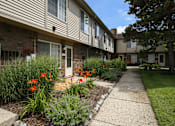 Thumbnail 4 of 47 - a sidewalk in front of an apartment building with flowers at Village Club of Rochester Hills, Shelby Township, MI 48317
