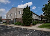 Thumbnail 9 of 47 - an apartment building in a parking lot with a sidewalk and a tree at Village Club of Rochester Hills, Michigan