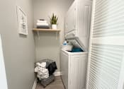 Thumbnail 20 of 35 - A small laundry room with a washer and dryer, a basket of clothes, and a shelf with a plant and towels.