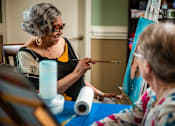Thumbnail 98 of 109 - An older woman is painting a picture while another woman looks on.