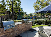 Thumbnail 8 of 57 - Apartments for Rent in Laguna Niguel CA - Pointe Niguel - BBQ Area with Two Grill Stations, Table, Chairs, Umbrella, Surrounding Trees, and View of Playground at Pointe Niguel, Laguna Niguel