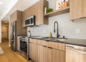 Thumbnail 4 of 30 - Two Bedroom Luxury Apartments in Downtown Oakland CA - Alice House - Modern Kitchen with Stainless Steel Appliances, Hexagonal Tile Backsplash, Wood-Style Cabinetry, Storage Shelving, and Hardwood Style Flooring