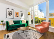 Thumbnail 9 of 30 - 2 Bedroom Luxury Apartments in Downtown Oakland CA - Alice House - Living Room with Hardwood Style Flooring, a Green Sofa, Salmon Armchair, Wooden Coffee Table, Striped Rug, Golden Side Table and Standing Lamp, White Walls, and a Large Window with Urban Views