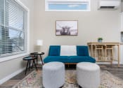 Thumbnail 29 of 97 - One-Bedroom Apartments in South Austin, TX - Casata Austin - Spacious Living Room with Wood-Style Flooring, Area Rug, Blue Couch, Round Ottomans, Side Tables with Lamp, Wooden Dining Table, AC Unit, Clerestory Window, and Large Window with Blinds.