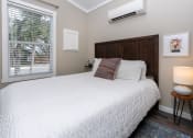Thumbnail 42 of 97 - 1-Bedroom Apartments in South Austin, TX - Casata Austin - Bedroom with Wood-Style Flooring, Area Rug, Bed with Wooden Headboard, Nightstand with Lamp, AC Unit, and Window with Blinds.