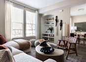 Thumbnail 23 of 33 - A living room with a white couch, a wooden chair, and a round table with a black object on it.