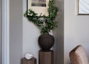 Thumbnail 27 of 33 - A plant in a pot sits on a pedestal next to a chair.
