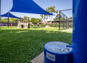Thumbnail 42 of 57 - A blue trash can with the label "Tool Dog" on it is in the foreground of a grassy area with a blue shade structure in the background.