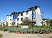 Thumbnail 1 of 87 - Dog-Friendly Apartments in West Sacramento, CA - The Strand - Exterior View of Apartment Complex with White Walls, Landscaped Plants, Trees, and Concrete Walking Path.