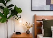 Thumbnail 59 of 85 - A modern bedroom with a wooden headboard and a side table with a lamp and a plant.