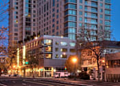 Thumbnail 1 of 104 - A modern multi-story building with glass windows is illuminated at dusk at The Grand Apartments, Oakland, CA