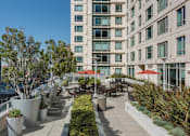 Thumbnail 102 of 104 - A patio area with tables and chairs is surrounded by a building and greenery at The Grand Apartments, Oakland, California
