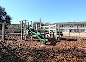 Thumbnail 1 of 7 - A playground with a green slide and brown leaves on the ground.