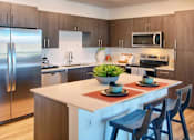 Thumbnail 16 of 35 - A modern kitchen with a stainless steel refrigerator, wooden cabinets, and a white island with a potted plant on top.