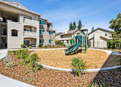 Thumbnail 11 of 28 - A playground with a green slide is surrounded by a white fence and brown mulch.