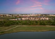 Thumbnail 33 of 33 - A large building complex is surrounded by a grassy field and a body of water under a pink and blue sky.