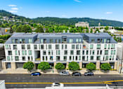 Thumbnail 34 of 35 - A large white building with a grey roof is surrounded by trees and cars.