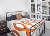 Thumbnail 10 of 35 - A bed with a black frame and an orange and white comforter is in the foreground of a room with a white wall and a window.