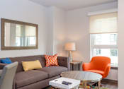 Thumbnail 3 of 7 - A living room with a grey couch, orange chair, and a coffee table.