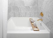 Thumbnail 20 of 35 - A white bathtub with a gold faucet and a tray with toiletries on it.