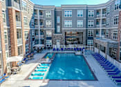 Thumbnail 2 of 23 - A large swimming pool surrounded by lounge chairs and umbrellas in a courtyard surrounded by apartment buildings.