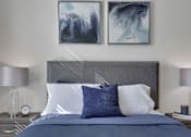 Thumbnail 18 of 23 - A bed with a blue comforter and two pillows with a grey headboard and two lamps on the side.