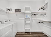 Thumbnail 30 of 43 - A white laundry room with a washer and dryer, a shelf with clothes, and a rack with clothes hangers.