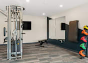 Thumbnail 36 of 98 - A gym room with a weight machine, a bench, and a mirror at The Beaumont Apartments, Bozeman