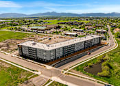 Thumbnail 82 of 98 - A large grey building is surrounded by a grassy area and a road with cars at The Beaumont Apartments, Bozeman, MT