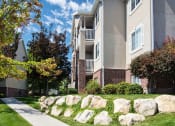 Thumbnail 1 of 5 - Side view of Country Oaks Apartment building with columns featuring part brick, large windows, white balcony railings, and pathway with rock landscaped yard area at Country Oaks in Clearfield, UT