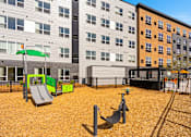 Thumbnail 62 of 98 - View of playground with a grey slide, play area, and two small swings with woodchips on the ground at The Beaumont Apartments, Bozeman, Montana 59715