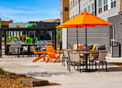 Thumbnail 66 of 98 - Outdoor resident patio area with a patio table and chairs with an orange umbrella and view of fire pit lounge with orange Adirondack chairs at The Beaumont Apartments, Montana